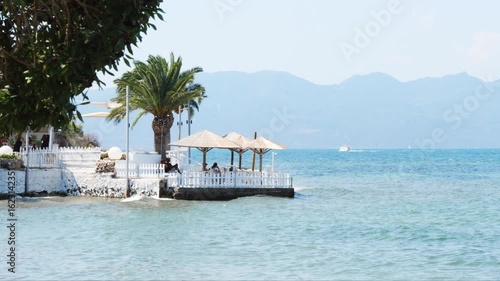 Wallpaper Mural Coastal café in Aegina, Greece with people sitting under straw umbrellas on a wooden platform above the sea. Palm tree nearby, white railing, lush greenery, calm blue water, distant boat and misty mou Torontodigital.ca
