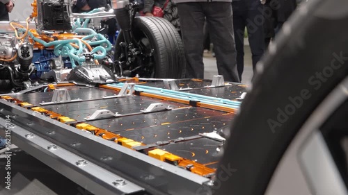 A group of people closely examining the chassis and powertrain of an electric vehicle (EV) at an auto show. The battery pack, orange high-voltage cables, suspension system, and electric motor.