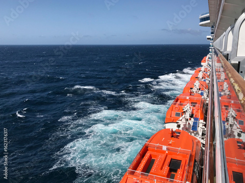 Orange lifeboats lined along the side of a cruise ship, viewed from a balcony with the deep blue ocean stretching out beyond, capturing both safety and serenity at sea.