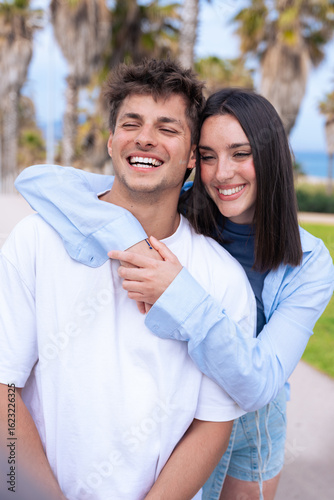 Vertical portrait Young couple embrace on a palm lined beach promenade, boyfriend laughing as girlfriend wraps arms around his shoulders from behind, capturing joyful love, travel vibes.