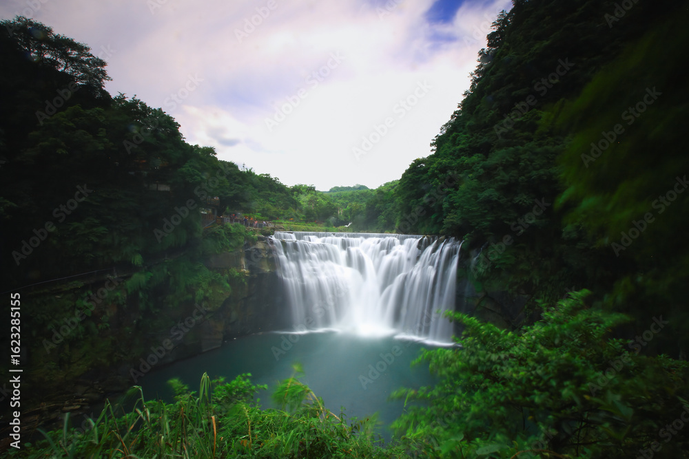 Fototapeta premium The breathtaking Shifen Waterfall plunges into an emerald pool, surrounded by lush green forests under a clear blue sky in Pingxi, New Taipei City, Taiwan, during summer.