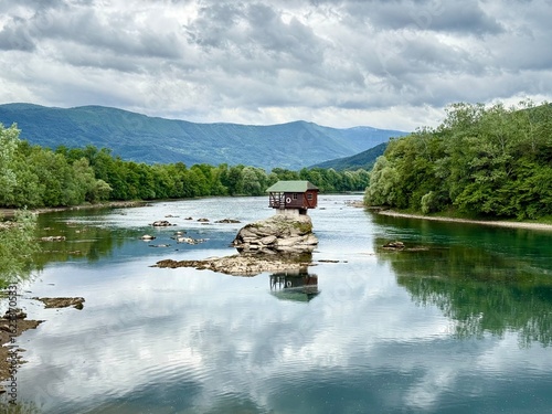 Drina river house in Serbia. A solitary wooden house stands on a rock in the middle of a calm river, surrounded by lush greenery and distant mountains. Serene forested area