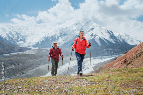 Schilderij op canvas Smiling backpackers couple with backpacks and trekking poles during Lenin peak ascent with mountain peaks in background