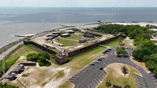 Aerial view of a historic fort surrounded by water, showcasing its large, star-shaped structure with multiple buildings and grassy areas. Nearby roads and parking areas are visible.