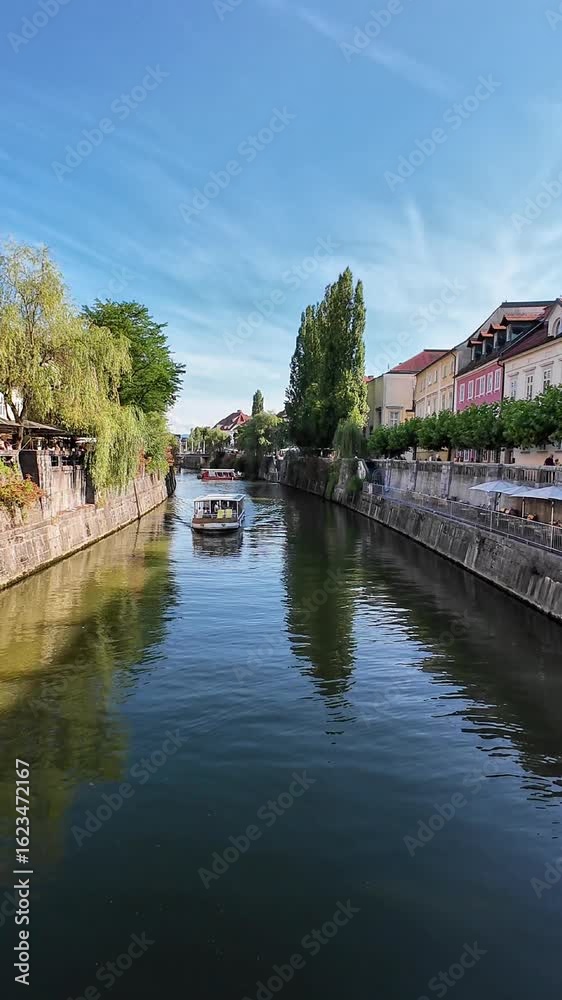 Tourist boat glides through Ljubljana’s canal, framed by historic buildings and lush riverside greenery Tourist boat glides through Ljubljana’s canal, framed by historic buildings and lush riverside greenery