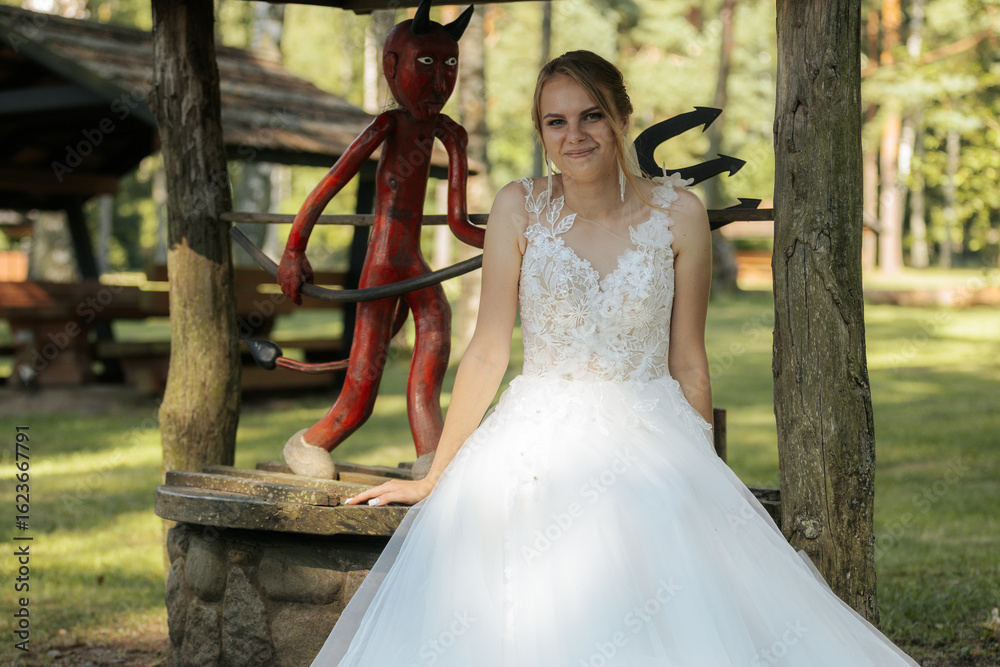 Naklejka premium A smiling bride in a white gown gestures toward a red devil sculpture at a rustic wooden well, blending humor and tradition in a whimsical wedding moment.