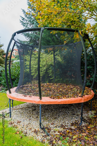 Vertical photo of a round trampoline in a garden, its surface covered with fallen autumn leaves. Surrounded by evergreen and maple trees, reflecting seasonal change and backyard leisure in fall.