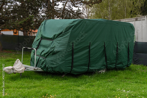 Travel trailer parked on grass and fully covered with green protective tarp and black straps. Outdoor RV storage protection in spring camping area.