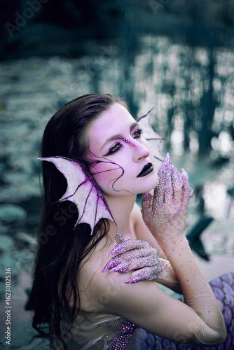 Mermaid sits in water with long hair and special makeup near lily pads during the evening light