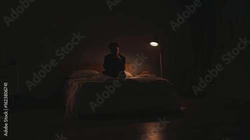 Young man sits on bed in dark room, illuminated by smartphone and floor lamp