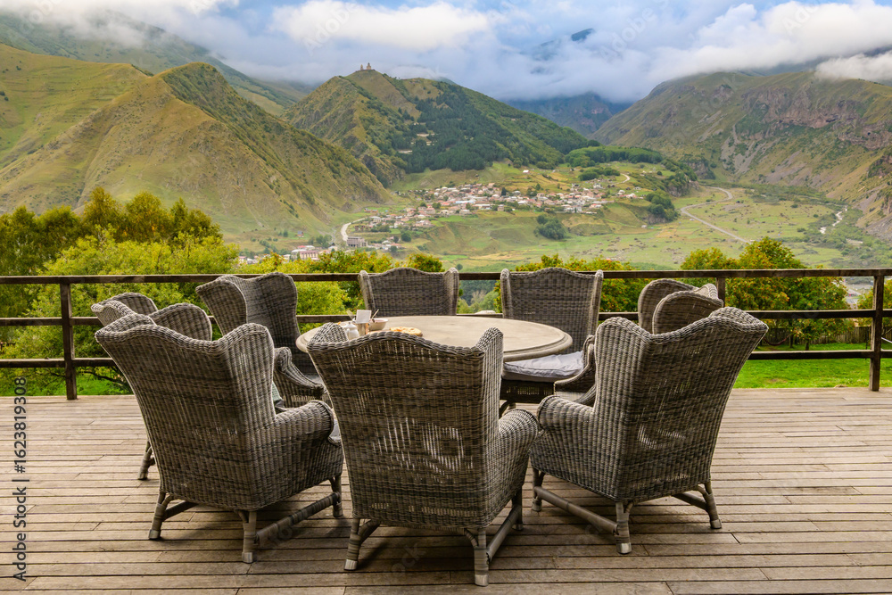 Fototapeta premium Elevated deck in Stepantsminda, with a view of the Caucasus and Gergeti Trinity Church. Wicker chairs surround a round table with refreshments.