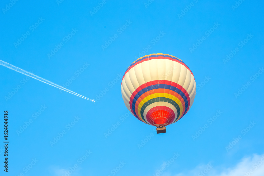 Obraz premium Colorful hot air balloon soaring high above clear blue sky with jet plane trail
