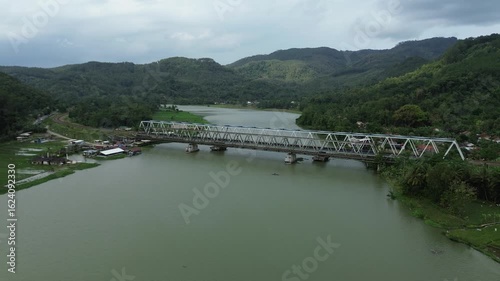 Wallpaper Mural Scenic aerial view of a steel truss bridge spanning a wide, calm river surrounded by lush green mountains and dense forest on a cloudy day. Torontodigital.ca