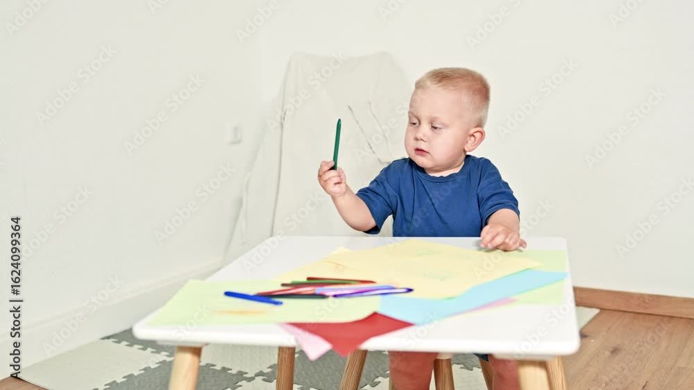 Cute 2 year old boy draws with colored pencils sitting at a children's table. Toddler is drawing at home. Child is developing and drawing pictures on paper. Boy in a T-shirt is engaged in creativity. Cute 2 year old boy draws with colored pencils sitting at a children's table. Toddler is drawing at home. Child is developing and drawing pictures on paper. Boy in a T-shirt is engaged in creativity.