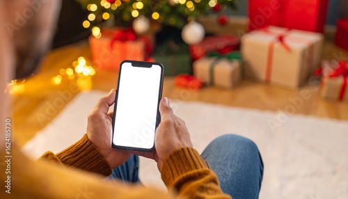 Person sits on floor, holding a phone with a blank screen, near a decorated Christmas tree and presents