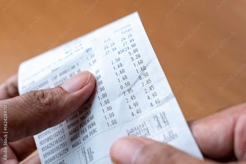 Fototapeta premium A person's hands are holding up and examining a long printed grocery receipt. Shopping, expenses, budgeting concept.