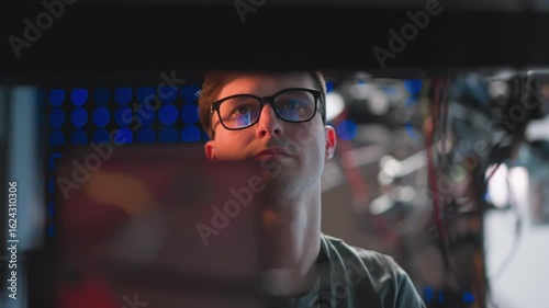 Close-up of focused person in glasses working at a computer, concentrating on tasks in a studio environment, illuminated by bright background lights, with reflections in glasses