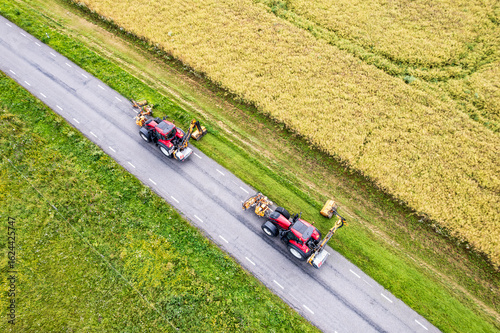 Drone view of two tractors with a mechanical mowers mowing grass on the side of the asphalt road