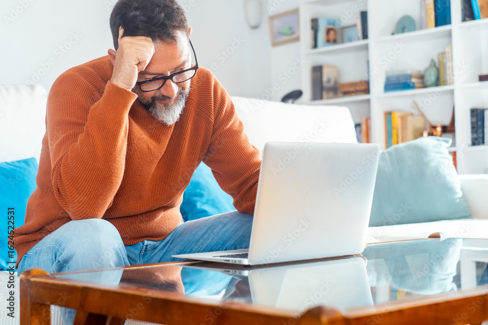 Obraz premium Middle-aged man working from home on laptop in living room, sitting on sofa with table, focused and stressed freelancer dealing with bills, household economy, deadlines and responsibilities
