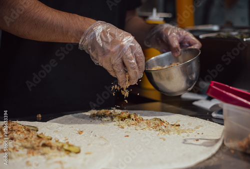 Dark atmospheric photo of cooking in a fast food shop. Chicken meat is boiled into pita bread. gloved hands of the cook create delicious street masterpieces of food. no face. table products and chef.