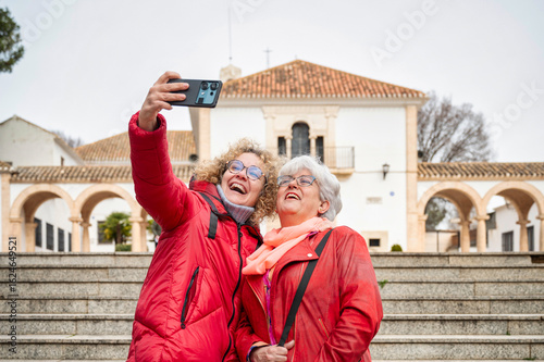Two mature women smiling while taking a selfie together during sightseeing trip, enjoying travel moments and digital memories in front of historical building