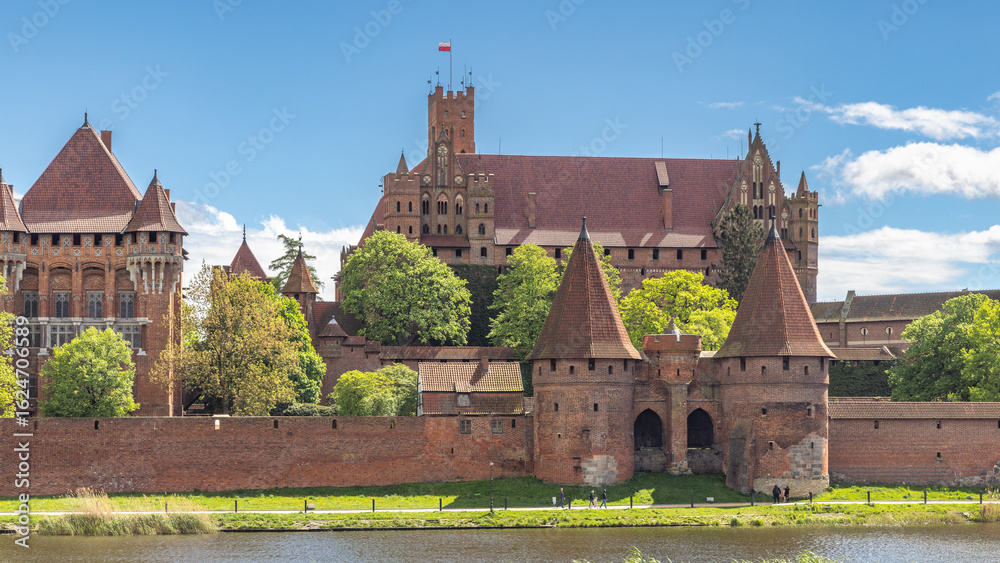 Naklejka premium Malbork Castle, a brick Gothic castle in northern Poland, Europe. Magnificent medieval castle, a testament to history and architecture, stands proudly against a backdrop of blue sky and lush greenery.