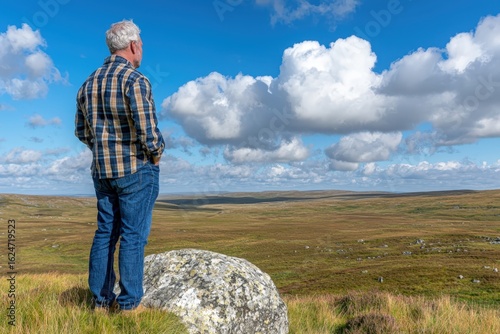 Man Stands on Rocky Outcrop Gazing at Expansive Countryside Under Bright Blue Sky with Fluffy Clouds in Beautiful Natural Landscape