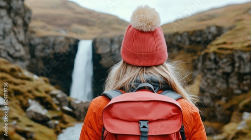 Young Adventurer in Red Hat and Backpack Stands Before Majestic Waterfall Surrounded by Lush Green Landscape in Stunning Wilderness Location