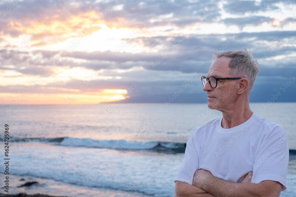 Fototapeta premium Senior mature man with crossed arms at the sea beach at sunset light looking the horizon over sea appreciating freedom and nature, handsome man enjoys a relaxed retirement lifestyle