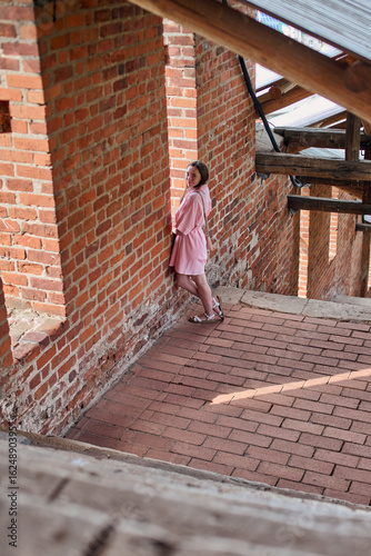 Young woman with brown hair in pink dress leans against red brick wall. She stands in sunlit alley under wooden beams, creating shadows and rustic atmosphere