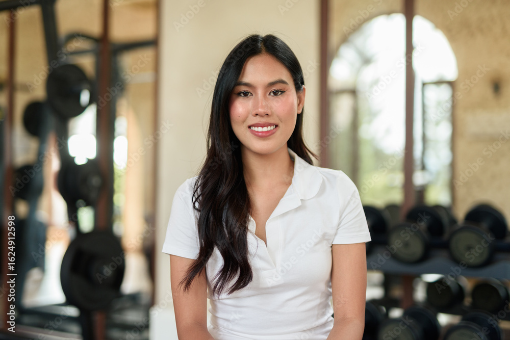 Fototapeta premium Young Asian woman in polo shirt at gym using phone in modern indoor training location