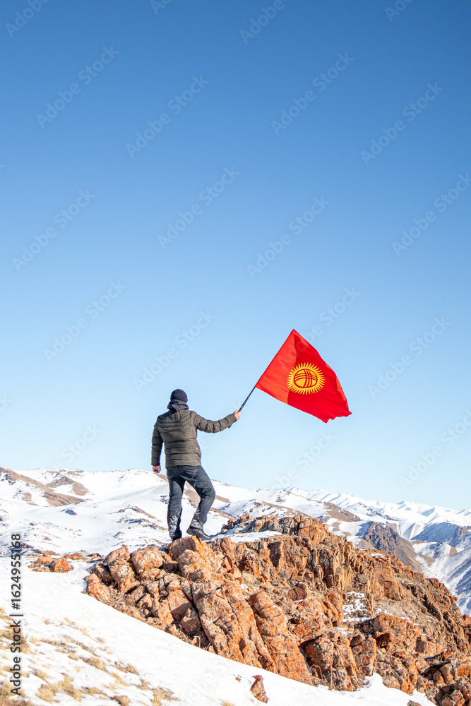 Obraz premium A man in winter clothing stands on a rocky mountain peak, holding a waving red flag of Kyrgyzstan with a vast snow-covered mountain range in the background