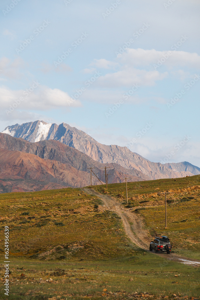 Fototapeta premium An off-road vehicle drives on a dirt road through a grassy valley, with majestic snowcapped mountains in the background under a blue sky