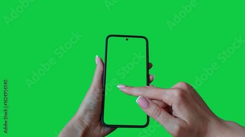 Young woman hand touch smartphone with green screen on green background. Phone in a female hand. Scrolling and swiping on a green phone screen. Template for scrolling through a feed in social network