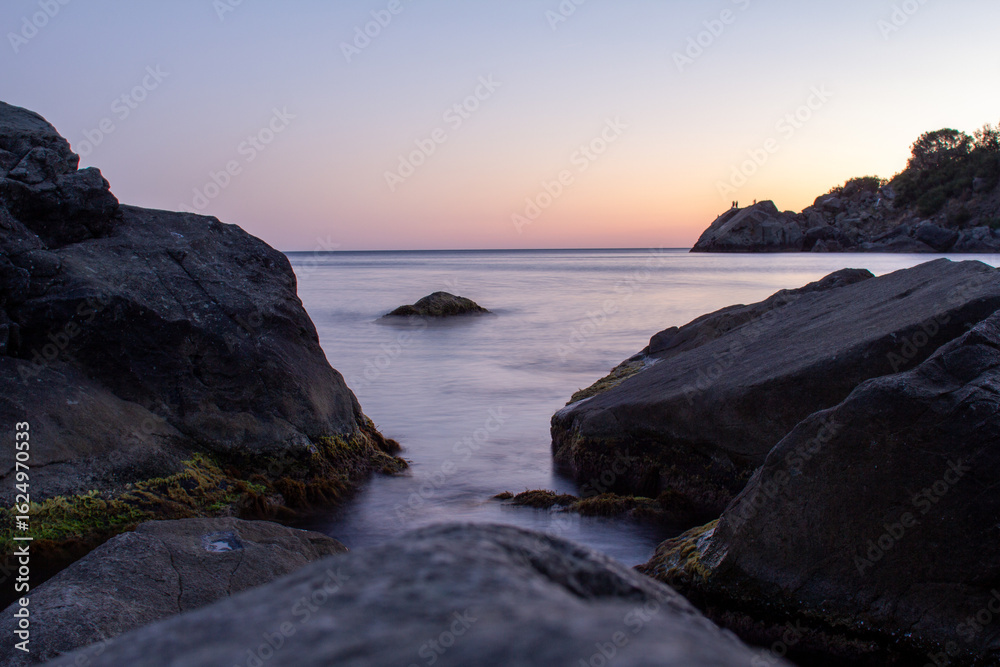 Fototapeta premium A stunning view of large, jagged rocks on a tranquil sea shore, with a small stream of calm water flowing between them, a perfect image for conveying themes of nature, coastal travel, or serene landsc