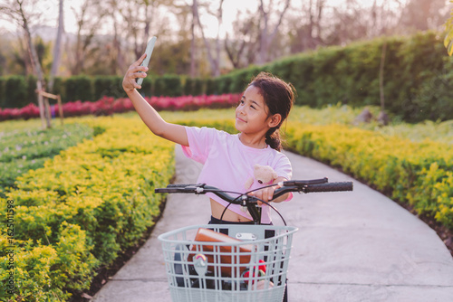 Cute Asian young girl doing activity outdoors in the nature park with her blue bicycle. She uses her mobile phone to take selfies and video calls.