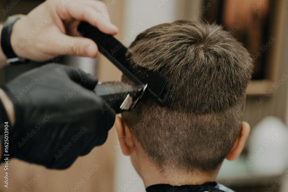 Fototapeta premium Smiling little boy sitting at barber shop, while hairdresser styling his hair.