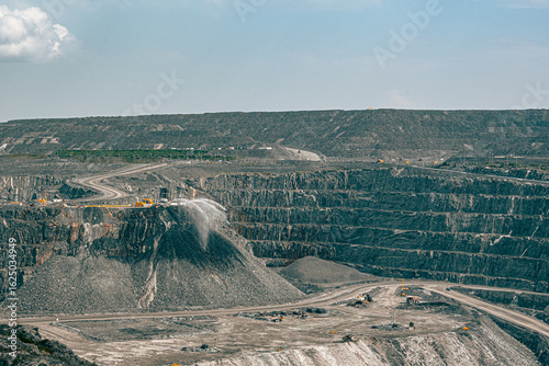 Wide view of a Canadian open-pit gold mine in Malartic, Quebec, showing heavy machinery and active dust control with water spraying on crushed rock. Industrial mining landscape.
