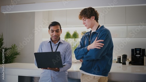 Two diverse people in modern startup office. thoughtful highschool student or intern gets mentorship while looking at laptop with colleague during project discussion
