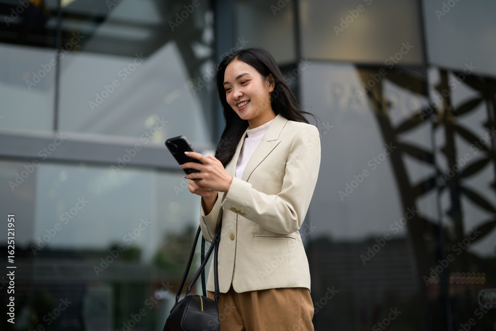 Fototapeta premium Smiling businesswoman using smartphone while walking near office building