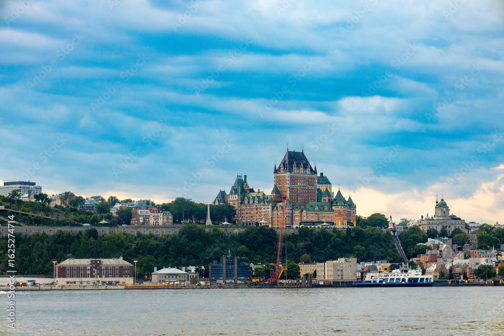 Obraz premium Distant view of Château Frontenac in Quebec City during sunset, with the St. Lawrence River in the foreground and warm light bathing the historic landmark.