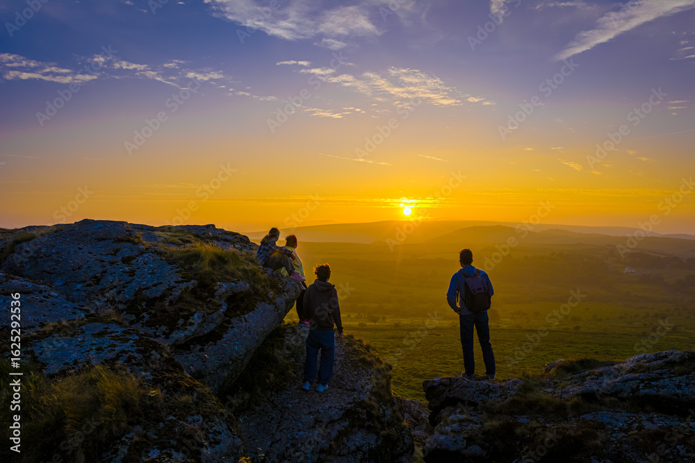 Family hiking on Dartmoor tors at sunset, silhouetted against dramatic skies in scenic moorland landscape Family hiking on Dartmoor tors at sunset, silhouetted against dramatic skies in scenic moorland landscape