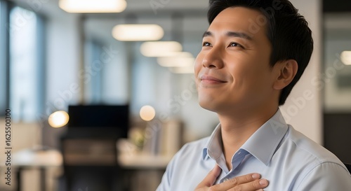A smiling Asian man in a light blue shirt, standing in an office with a blurred background, looking away from the camera with a thoughtful expression.