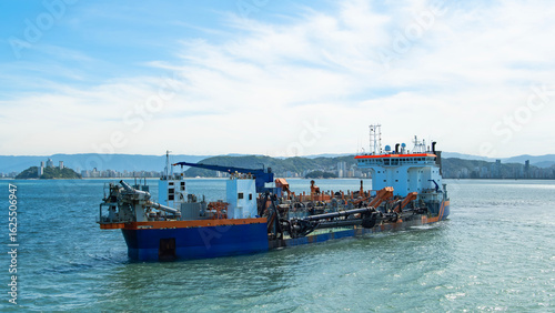 Large dredging vessel operating in coastal waters near a cityscape and mountains, equipped with pipes and cranes for marine construction work