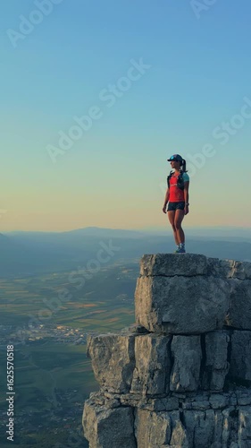 Determined female runner triumphantly stands atop a rugged cliff at sunrise, overlooking a vast valley. Clad in athletic gear, she embodies endurance and achievement.