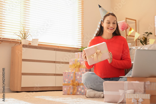 Young woman in party hat with gift and laptop celebrating Birthday online at ...
