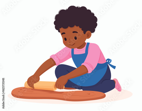 A curly-haired boy, wearing a dark blue apron over a pink sweater, sits on the floor and rolls a lump of terracotta-colored clay into the shape of a long snake.