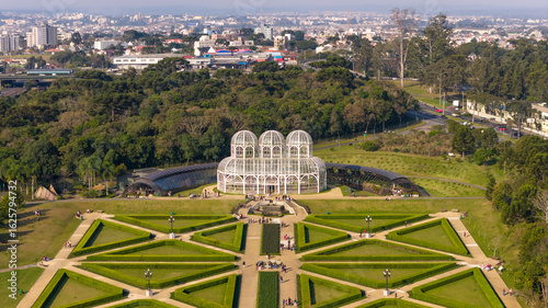 Front aerial shot of Curitiba’s iconic greenhouse and formal gardens with surrounding forest and distant city.