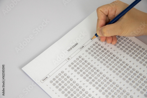 The image shows a hand holding a pencil and filling in a multiple-choice answer sheet for a test. The paper has a grid of circles
