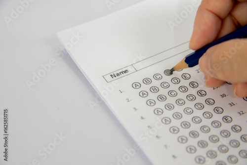 The image shows a hand holding a pencil and filling in a multiple-choice answer sheet for a test. The paper has a grid of circles